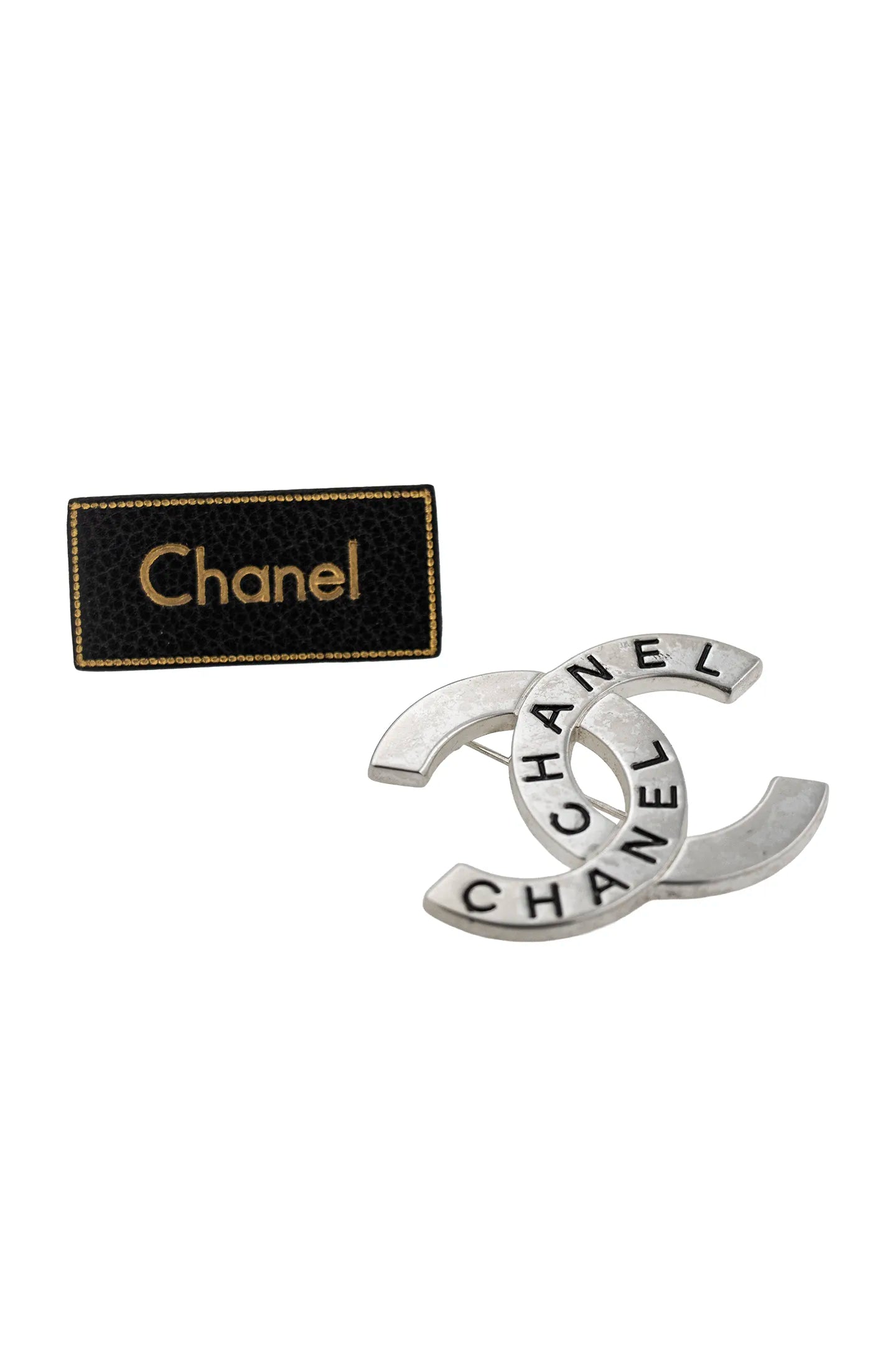 Étiquette cuir noir et doré Chanel posée près d'une broche CC argentée gravée du mot Chanel en relief.